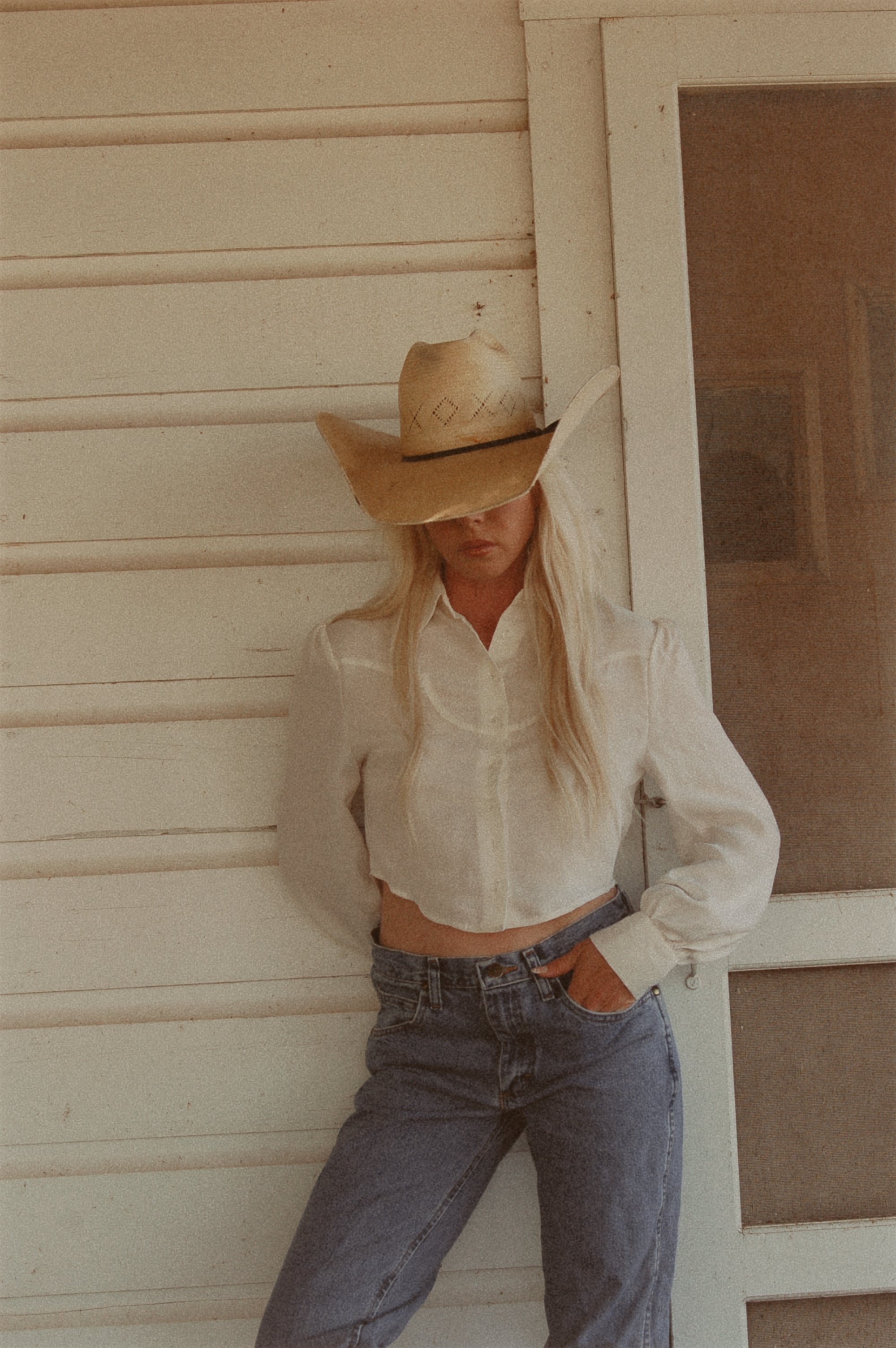 Person wearing a cowboy hat and white shirt leaning against a wall.